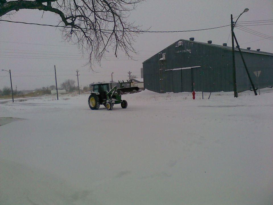 Green tractor with front loader in snow, next to a large building