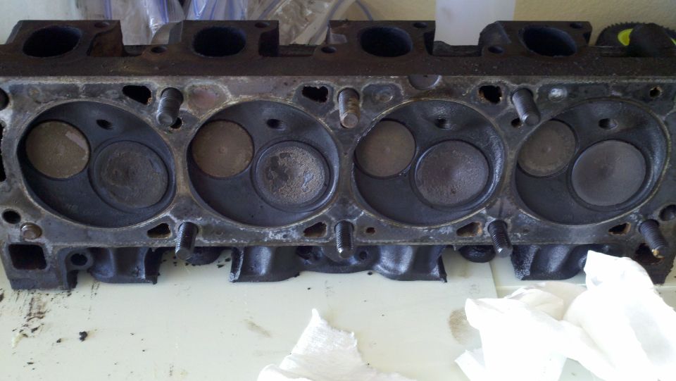 Engine cylinder head with four visible combustion chambers and valves, resting on a white surface
