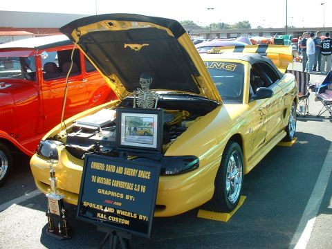 1995 Ford Crown Victoria - A yellow 1998 Ford Mustang convertible is displayed with its hood open, a skeleton and a sign listing owners and custom parts.