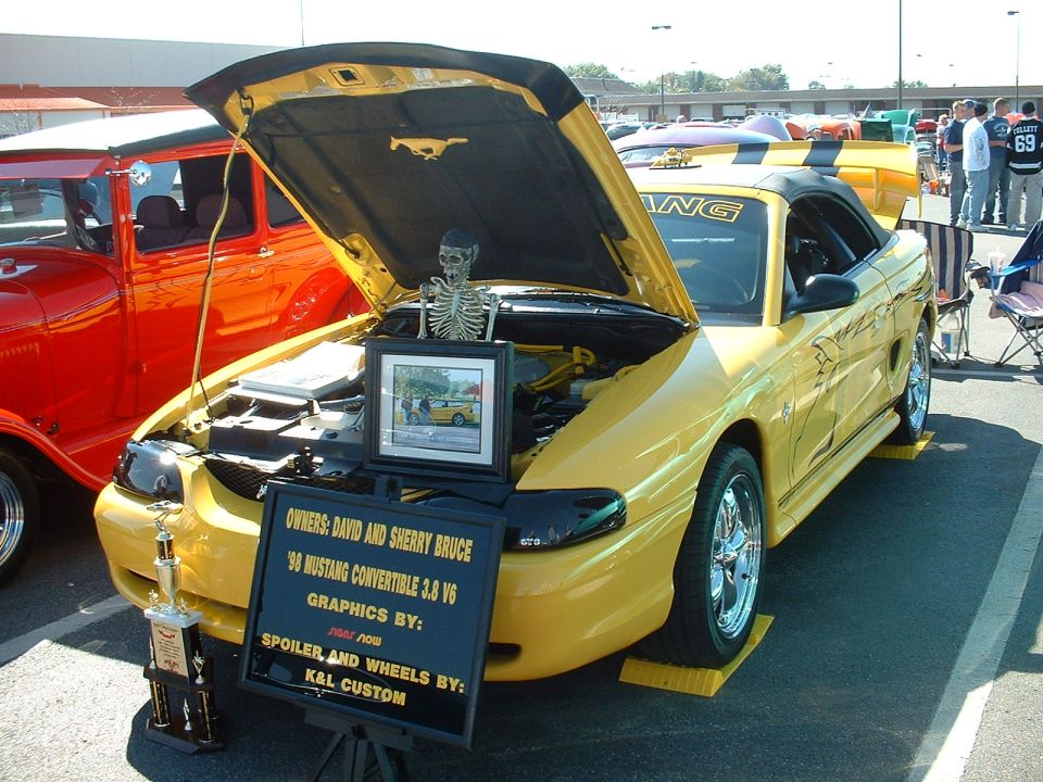 Yellow 1998 Ford Mustang convertible with hood open, displaying a skeleton and a sign detailing owners and modifications
