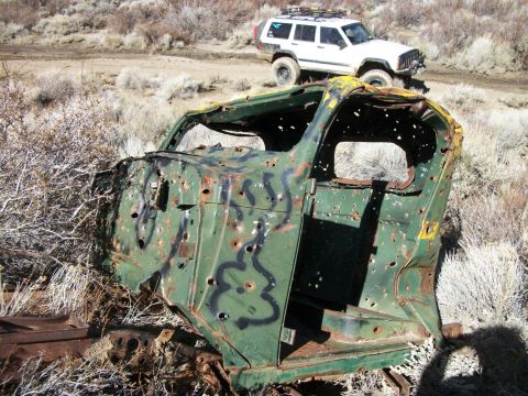 1996 Ford Bronco - A heavily damaged and bullet-riddled vehicle cab sits in a dry, brushy landscape with a white SUV in the background.