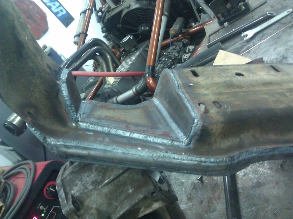 Welded metal frame section with engine components and exhaust pipes in background