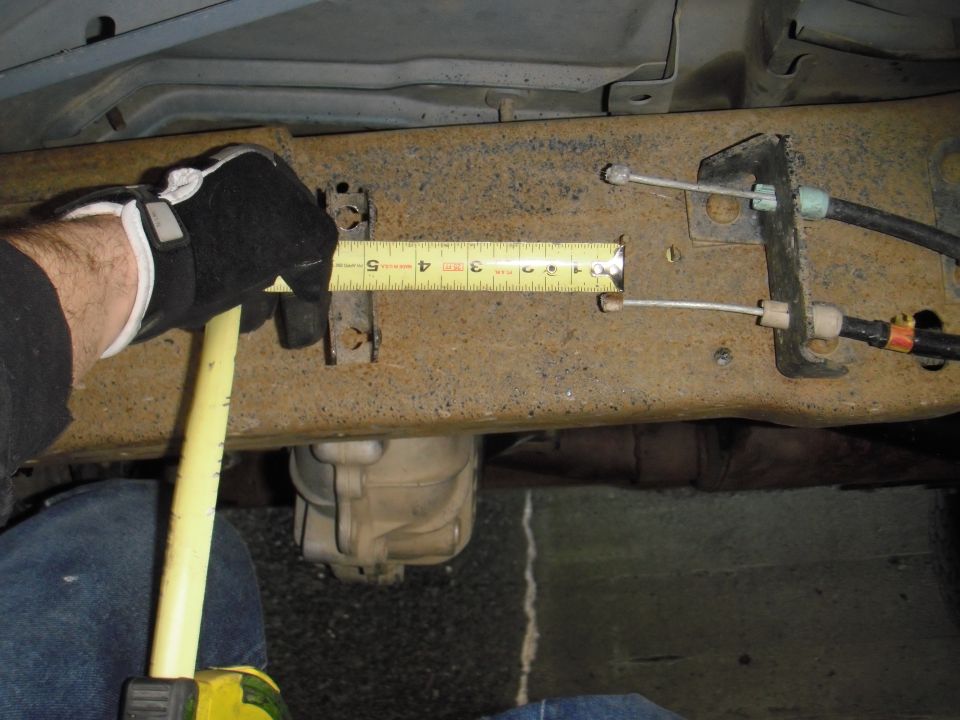 Gloved hand holding tape measure against rusty vehicle frame, measuring a section near brake cables