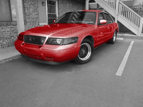 2000 Mercury Grand Marquis - A red 2000 Mercury Grand Marquis is parked in a lot with a brick building and stairs in the background.