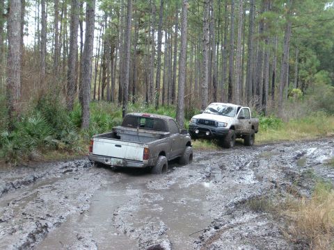 1991 Ford Bronco - Two pickup trucks are stuck in deep mud on a wooded trail, with one vehicle partially submerged.