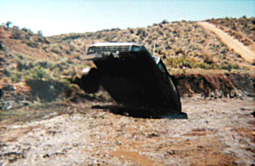 1981 Ford Bronco - A black SUV lies overturned on a dusty, rural road, with a dirt track visible in the background.