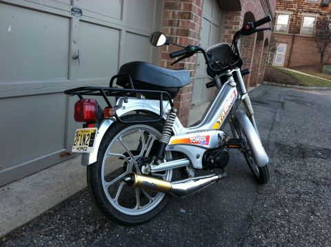 1999 Tomos Targa - A silver Tomos moped with a gold exhaust pipe is parked in front of a garage door.