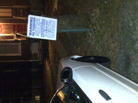 2000 Ford Crown Victoria - A white car is parked near a sign warning against parking, with a portion of its body and side mirror visible.