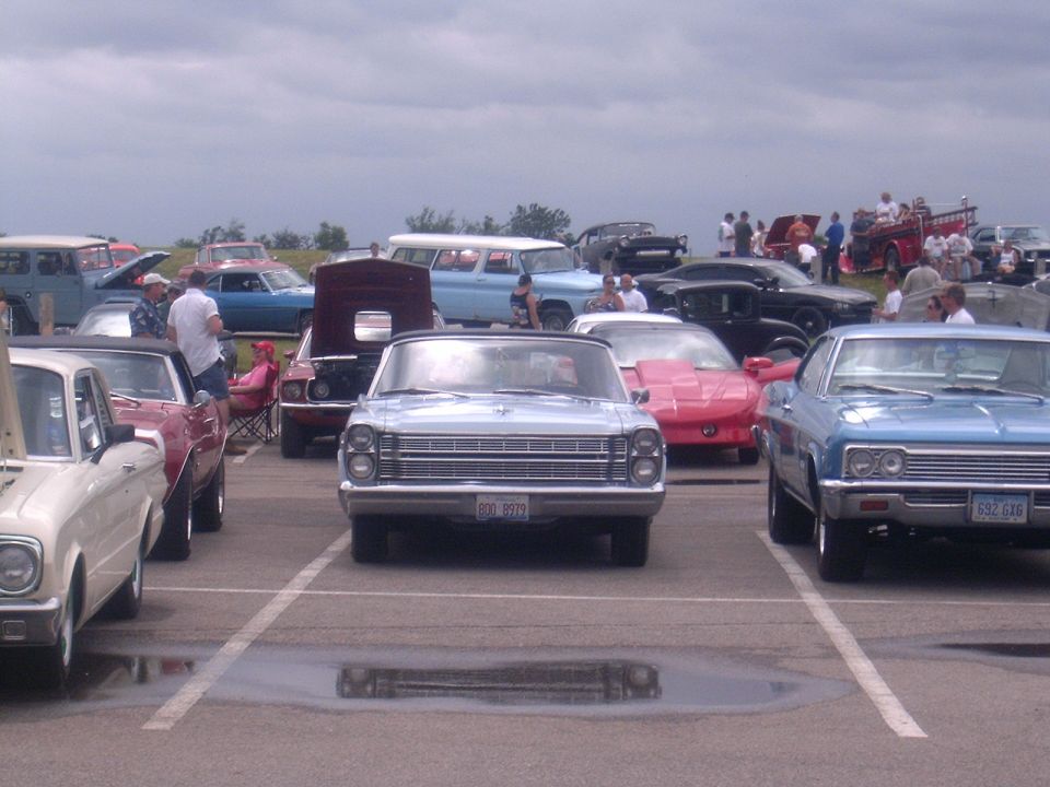 Classic car show with a silver Ford sedan front and center among vintage cars in a lot