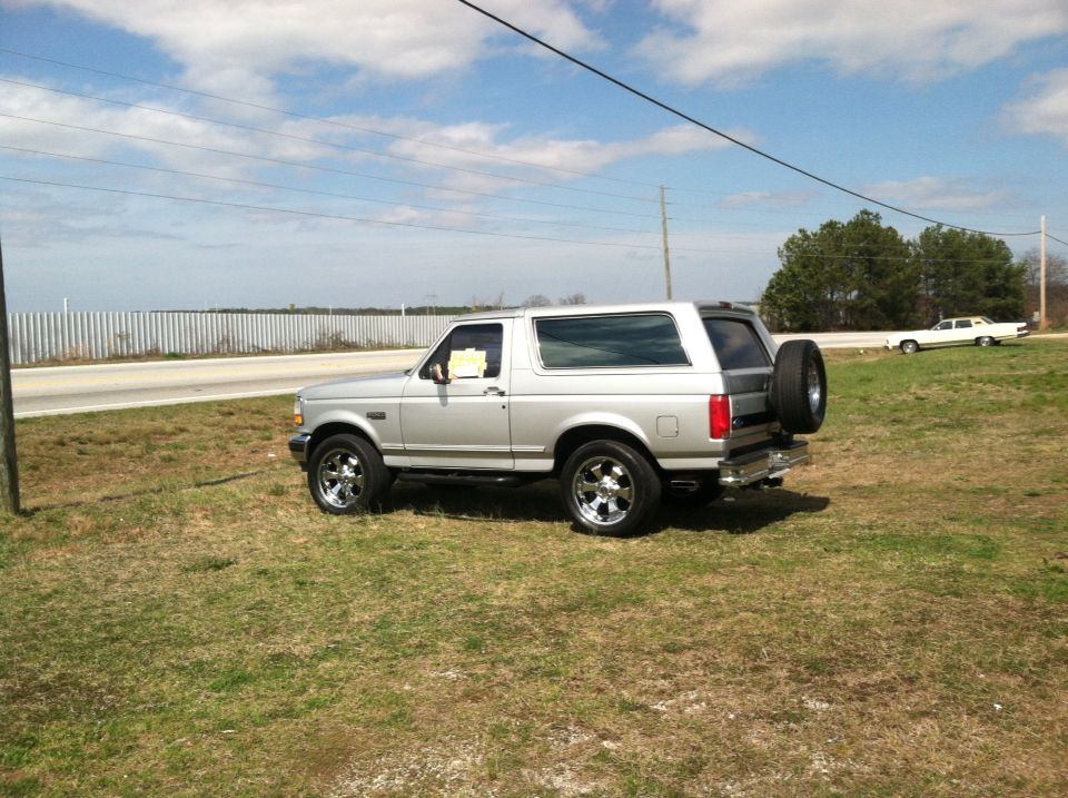 Silver 1993 Ford Bronco parked on grass next to a road, with another car in the distance