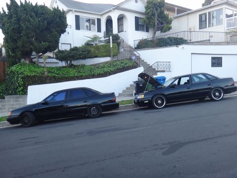 1993 Ford Taurus - Two black Ford Taurus sedans are parked on a street in front of a house, with one car's hood open.