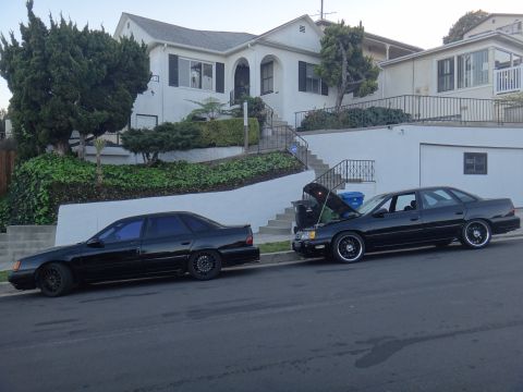 1993 Ford Taurus - Two black sedans are parked on a residential street, one with its hood open, suggesting recent maintenance or a problem.