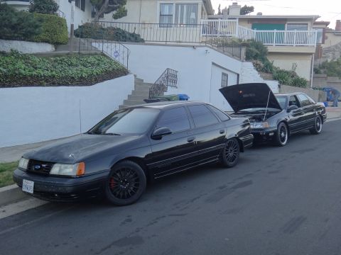 1993 Ford Taurus - Two black sedans are parked on a residential street, one with its hood up.
