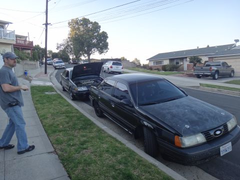 1993 Ford Taurus - A black sedan is parked on the street with its hood slightly ajar, while another car with its hood open is behind it.