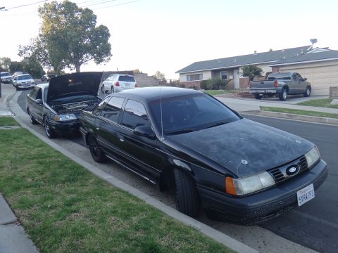 1993 Ford Taurus - A black 1993 Ford Taurus is parked on the street, with another car behind it having its hood open.