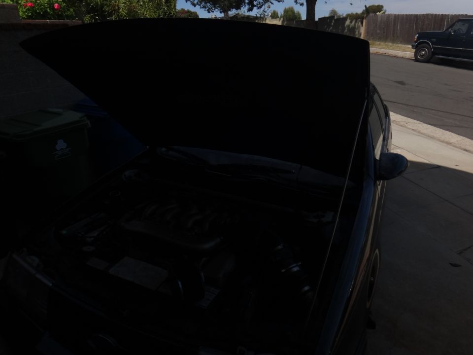 Car hood open showing the engine bay. A trash can is to the left, and a dark pickup truck is parked on the street in the background