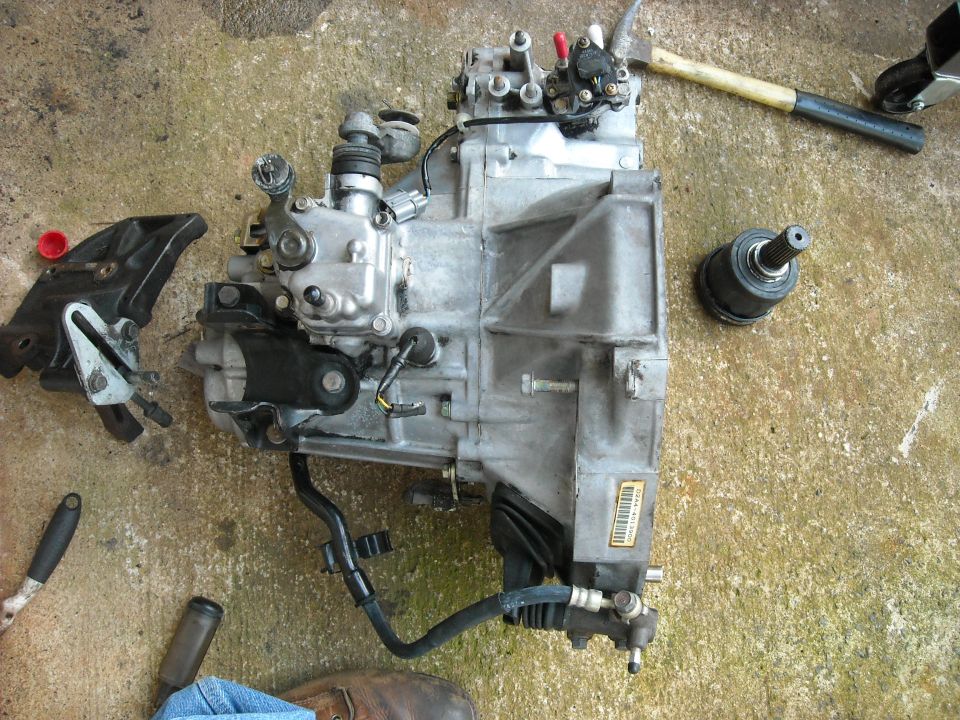 car transmission with attached hoses and mounting bracket, lying on concrete floor next to a CV joint