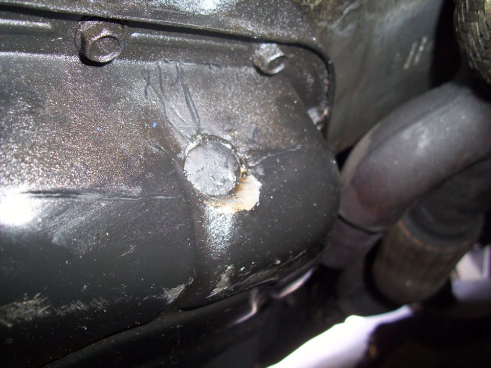 Black engine oil pan with a circular drain plug opening and some sealant residue. Exhaust pipes are visible to the right