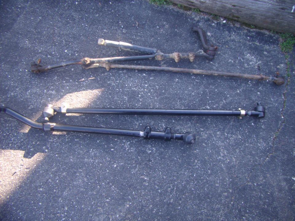Two sets of steering and suspension rods laid out on asphalt. One set is rusty and aged, the other is black and appears newer
