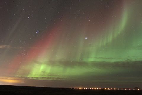 1970 Ford Mustang - The aurora borealis displays vibrant red and green hues across the night sky, with a line of city lights visible on the horizon.