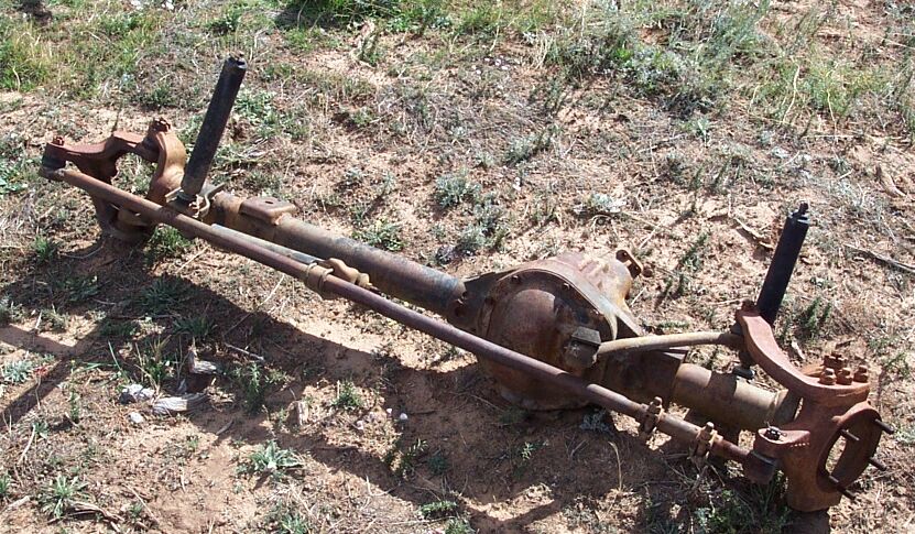 Rusty solid front axle with differential, tie rod, and shocks lying on dirt ground