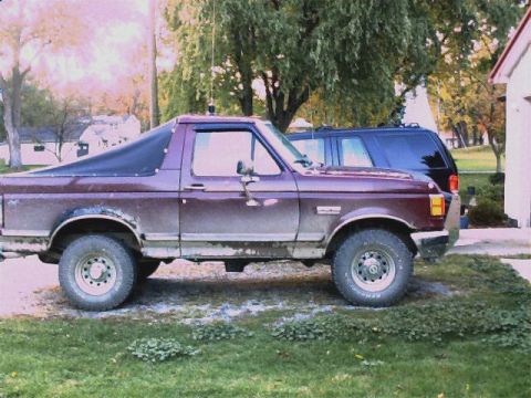1990 Ford Bronco - This maroon Ford Bronco, seen from the side, has a black soft top and visible rust on its lower body panels.