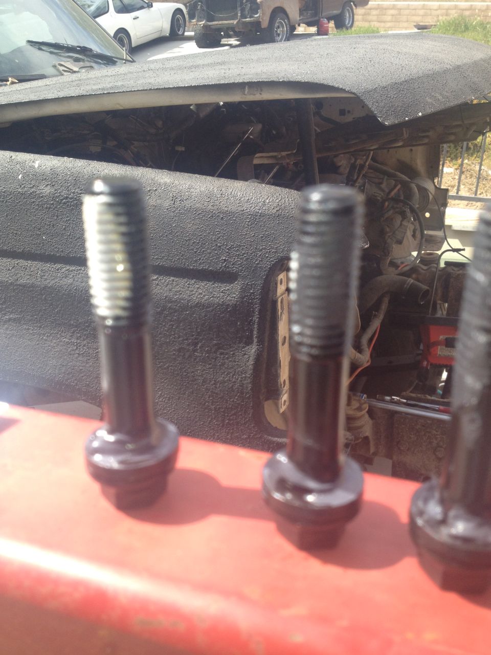 Three black bolts in focus, with an engine bay and other vehicles blurred in the background
