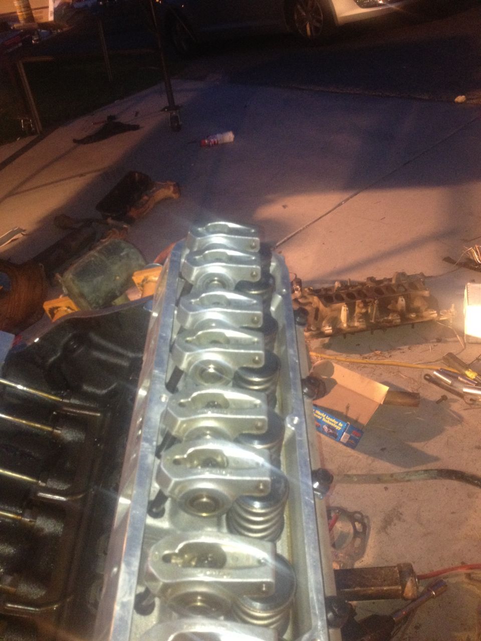 Close-up of an engine cylinder head with rocker arms and springs visible. Other engine parts and tools are scattered on the ground in the background