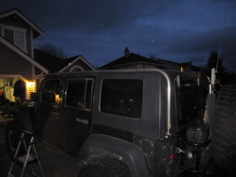 2008 Jeep Wrangler - A dark SUV with a roof rack is parked outside a house at dusk. The spare tire is mounted on the rear.
