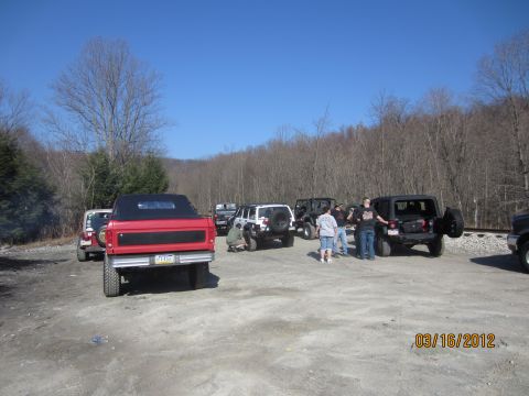 1979 Ford Bronco - Several off-road vehicles are gathered in a dirt lot with trees in the background.