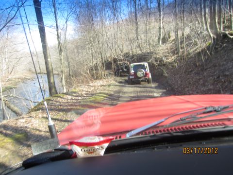 1979 Ford Bronco - Two Jeeps navigate a wooded dirt road next to a river, viewed from the interior of a red vehicle.