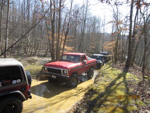 1979 Ford Bronco - A convoy of off-road vehicles navigates a wet, wooded path on a sunny day.