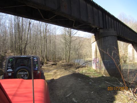 1979 Ford Bronco - Two Jeeps navigate a muddy path beneath a weathered railway bridge, with a river visible to the right.