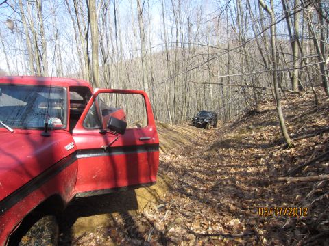 1979 Ford Bronco - Two off-road vehicles navigate a dirt path through a forest with bare trees.