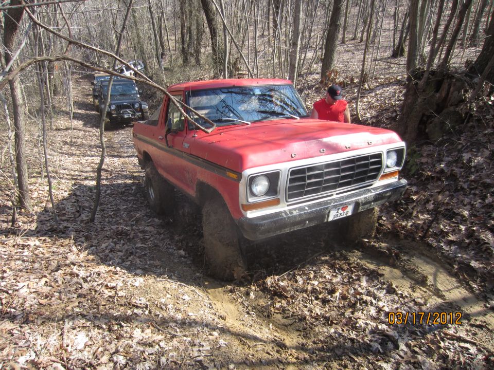 Red SUV driving on a muddy trail in woods. Another SUV is behind it