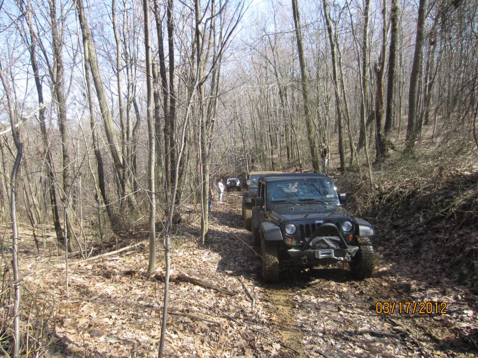 Black Jeep Wrangler on a muddy forest trail, with other vehicles behind