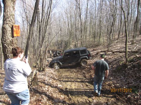 1979 Ford Bronco - Two people watch a Jeep Wrangler navigate a wooded off-road trail with another Jeep visible in the background.