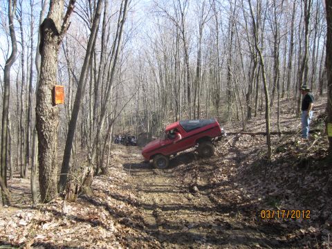 1979 Ford Bronco - A red 1978 Ford Bronco navigates a challenging muddy incline in a wooded area, with other off-road vehicles visible in the background.