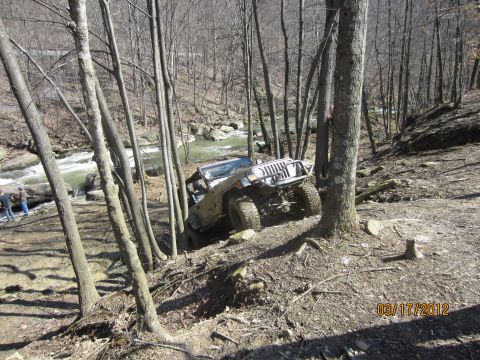 1979 Ford Bronco - A Jeep Wrangler tackles a challenging off-road course near a flowing river, surrounded by trees.