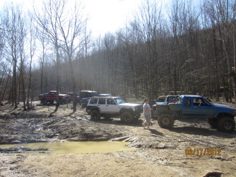 1979 Ford Bronco - Several off-road vehicles are parked in a muddy, wooded area on a sunny day.