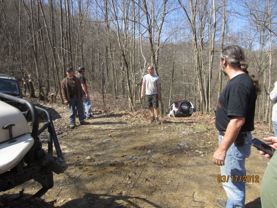 Several people stand around off-road vehicles in a wooded area; one vehicle is overturned