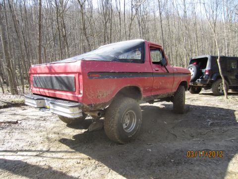 1979 Ford Bronco - A red 1979 Ford Bronco is seen from the rear 3/4 angle, equipped with off-road tires and a black soft top, parked in a wooded area.