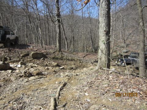 1979 Ford Bronco - Two off-road vehicles navigate a challenging, rocky path through a dense forest on a clear day.