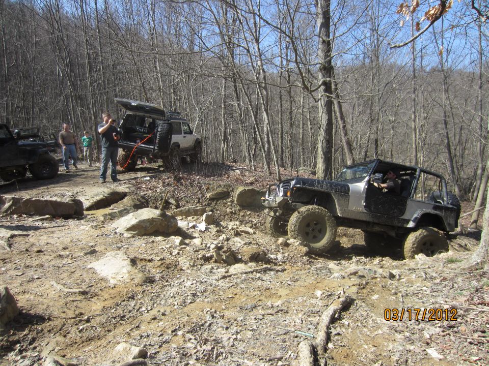 Several Jeeps navigating a rocky, wooded trail. One Jeep is in the foreground, angled to the right