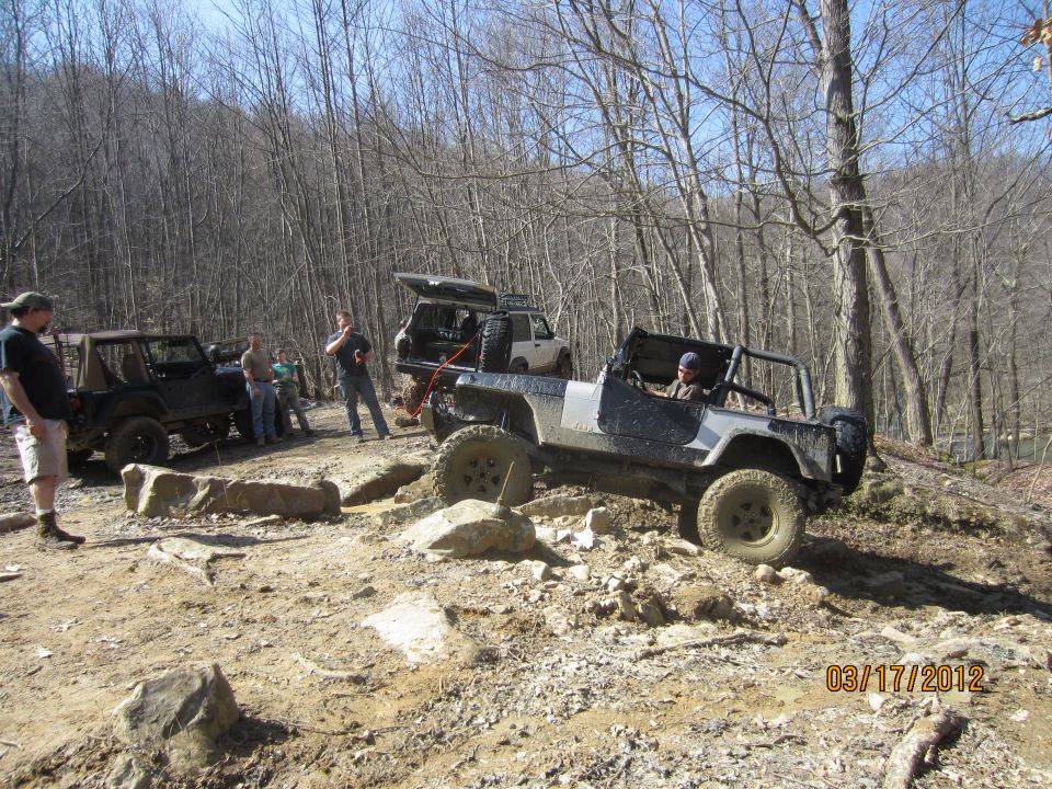 Several off-road vehicles are navigating a rocky, wooded trail. One Jeep is in the foreground, going over rocks