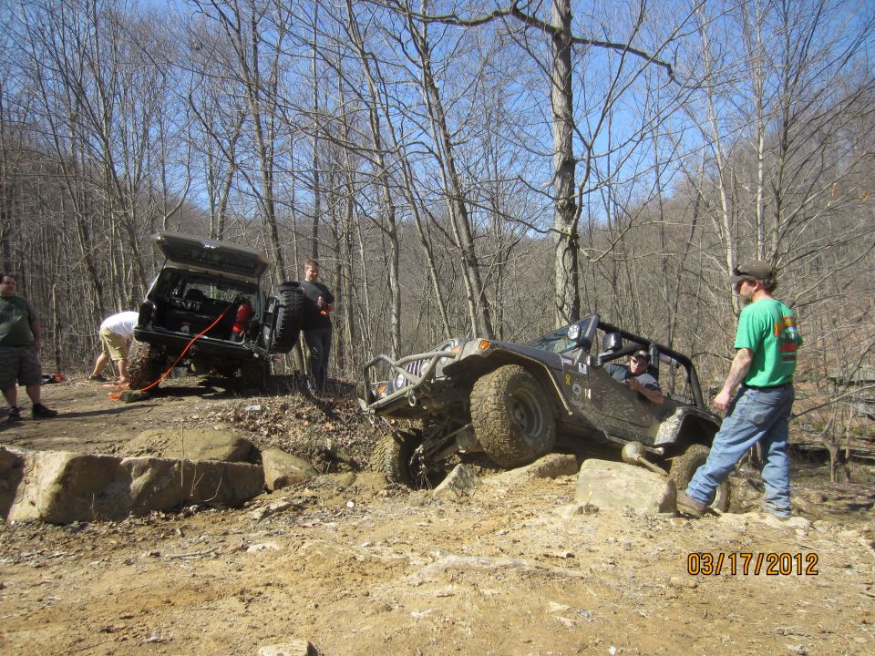Two off-road vehicles are on a dirt trail with trees around. One vehicle is on its side, and people are working on it. The other vehicle is in the foreground, also on a rocky path