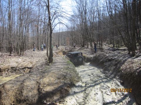 1979 Ford Bronco - A black Jeep Wrangler tackles a challenging muddy path surrounded by trees, with spectators watching from the side.