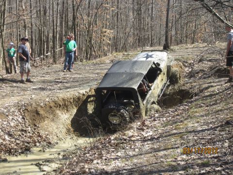 1979 Ford Bronco - Off-road vehicle stuck in a muddy ditch on a wooded trail, with spectators watching.