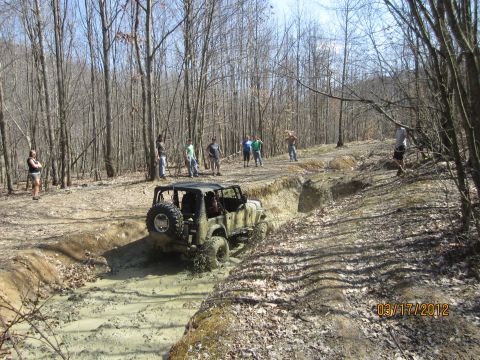 1979 Ford Bronco - A Jeep Wrangler drives through a muddy obstacle course in a forest with spectators watching.