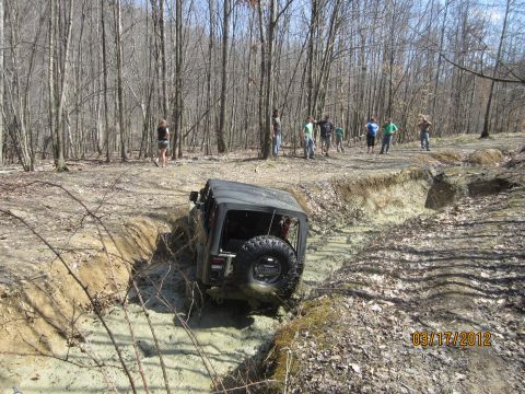 1979 Ford Bronco - A group of people watch as a black Jeep Wrangler tackles a challenging muddy obstacle on an off-road trail.